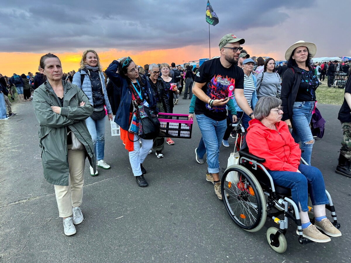 A group of about fifteen people moves across the festival area. A woman in a wheelchair is in the foreground. The background shows sunset light and a large number of participants.